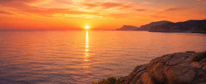 Mediterranean sunset over a calm sea with a rocky coastline and warm orange sky